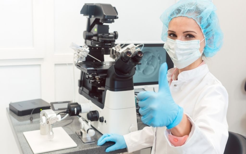 an embryologist showing thumbs up & seeing at camera and other embryologists equipments near her.